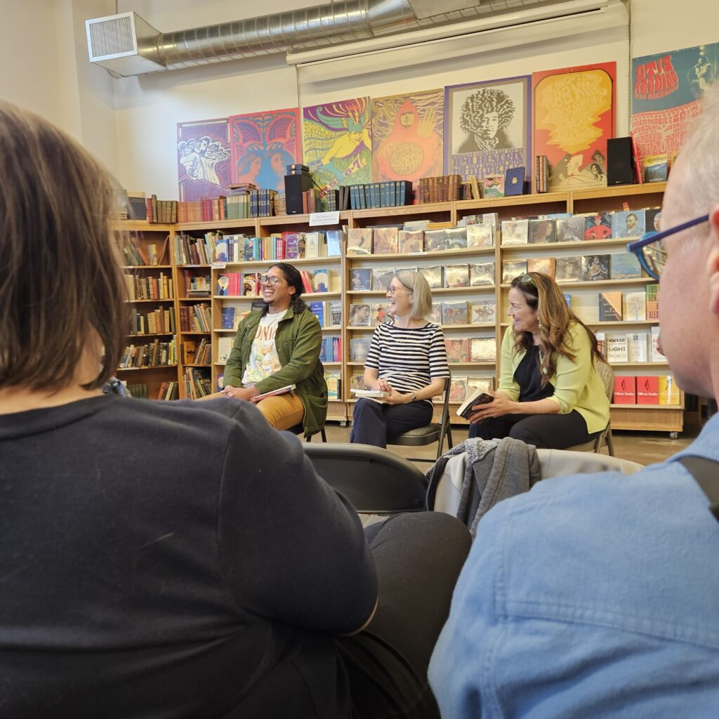 Three Sacramento Noir authors speak to an audience inside Beers Books on S Street in Sacramento, seated in front of bookshelves with colorful posters behind them.