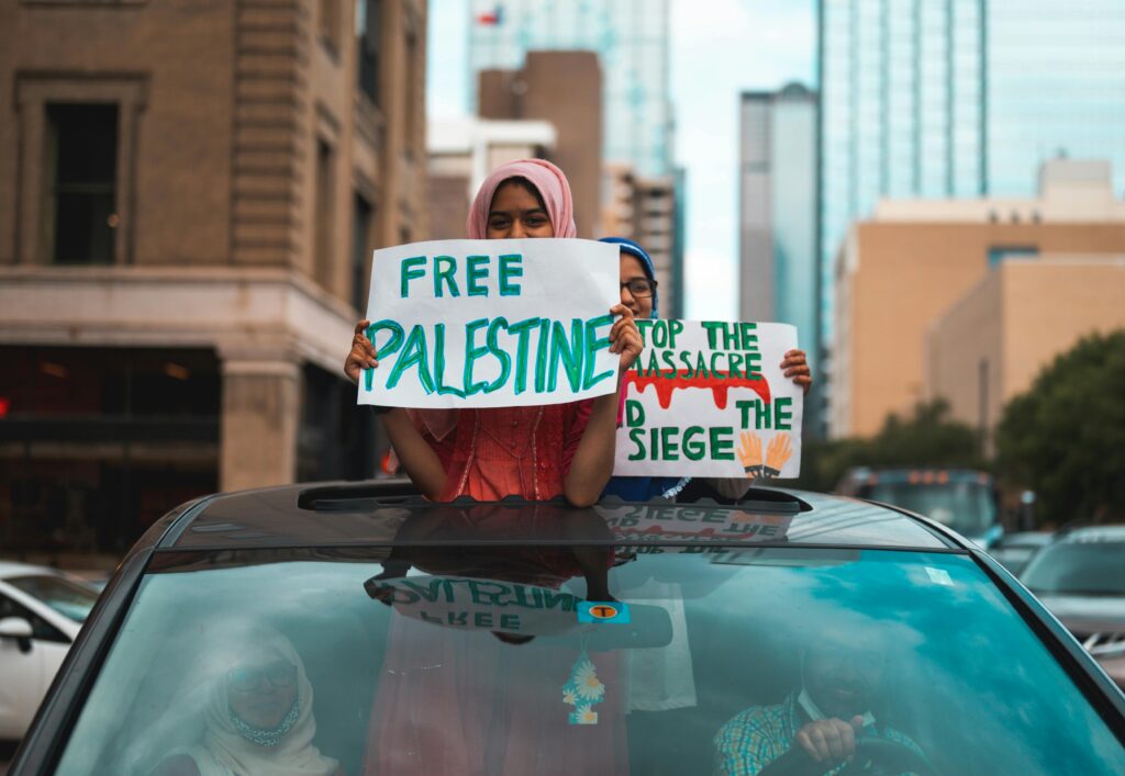 Two young protesters stand through a car’s sunroof holding handmade signs that read “Free Palestine” and “Stop the Massacre, End the Siege,” during a pro-Palestine demonstration in an urban downtown area. Photo by Alfo Medeiros via Pexels.