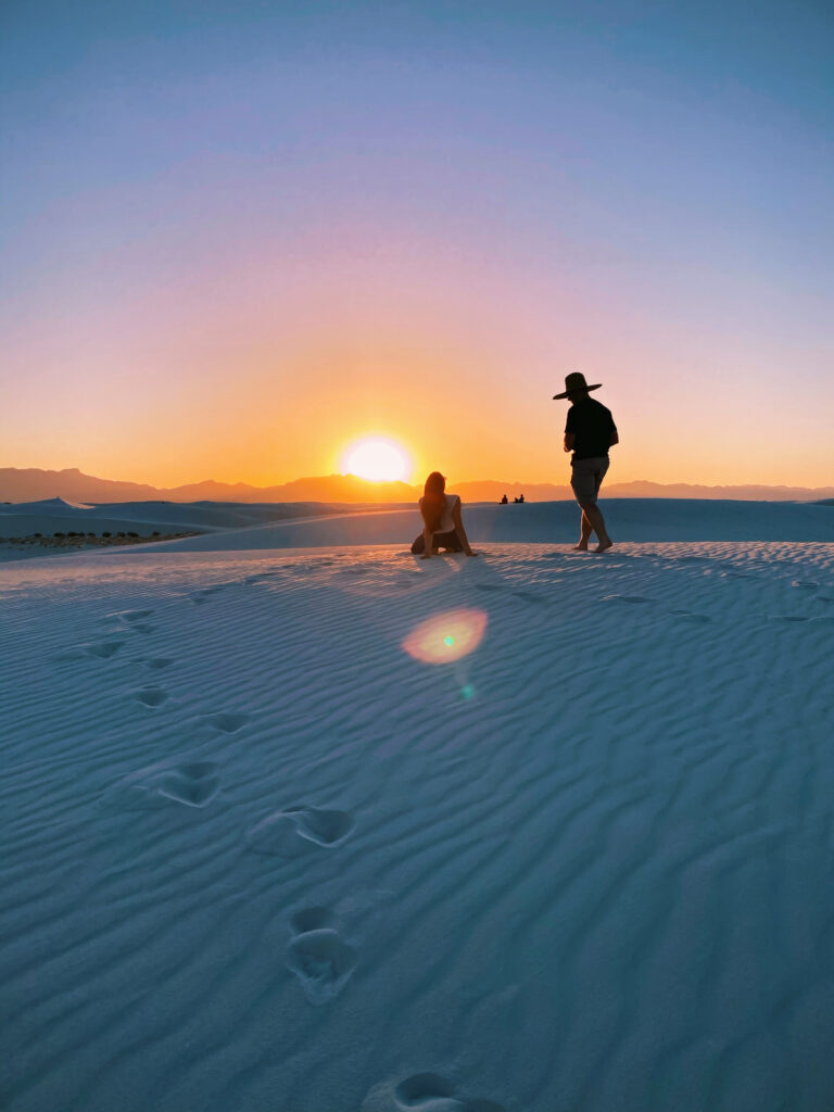 Two people standing and sitting on white sand dunes at sunset, with footprints leading toward the horizon and golden light in the sky.