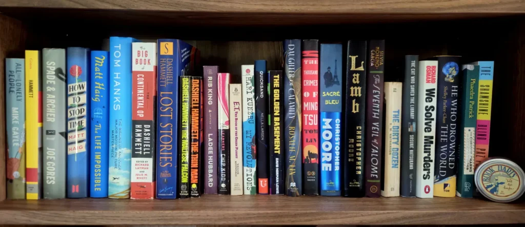 A full home bookshelf with a colorful row of books, including Tom Hanks, Matt Haig’s The Life Impossible, several Dashiell Hammett titles, and We Solve Murders, with a small “Book Darts” tin at the right end.